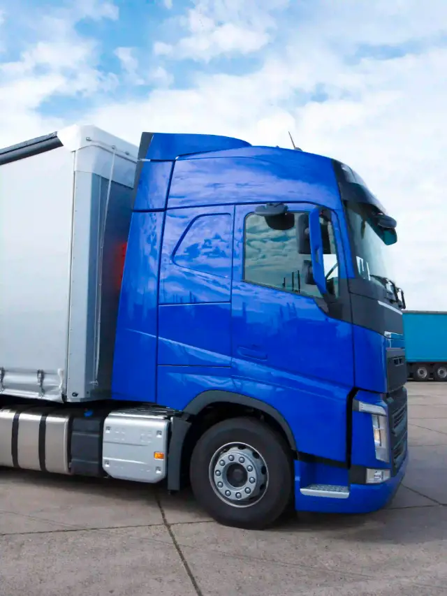 Cabine de profil d'un camion Volvo bleu avec remorque blanche stationné sous un ciel nuageux.