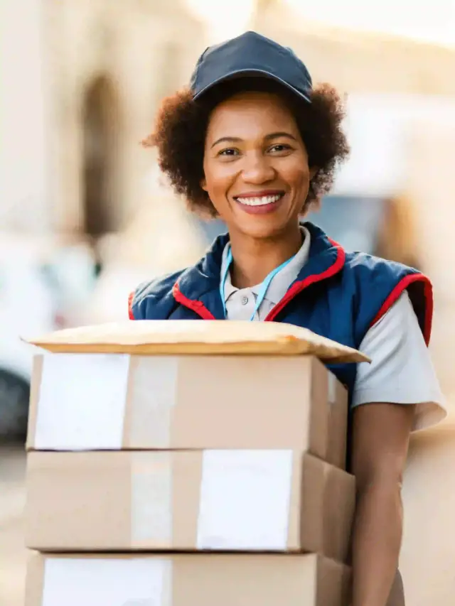 Une livreuse souriante portant une pile de colis en carton et une enveloppe dans la rue.